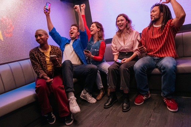 A young person with curly hair smiles at the camera while sitting in front of a laptop against a dark background.