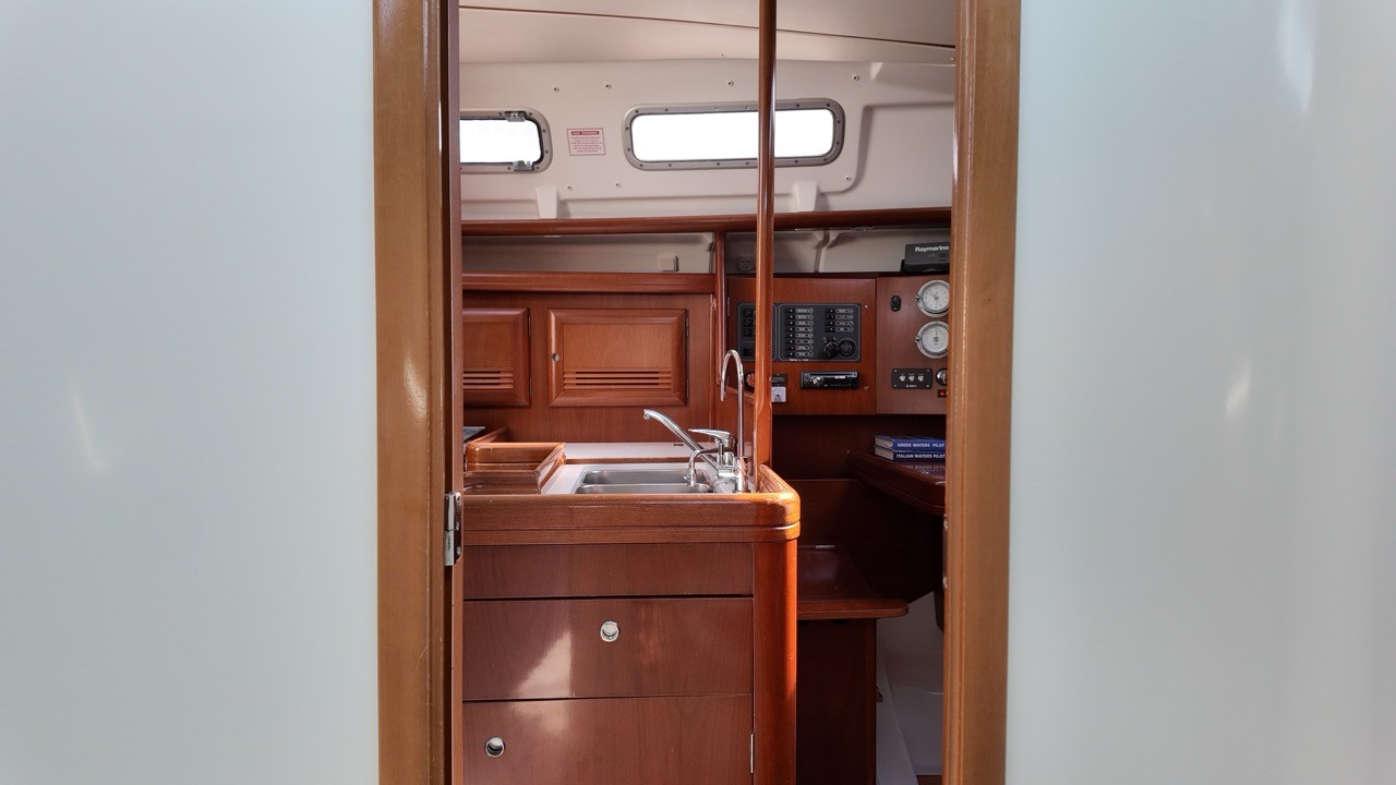 View through the companionway hatch of a Beneteau Oceanis Clipper 373 looking down into the saloon, revealing the teak-finish galley with stainless steel sink, multiple storage drawers, Raymarine electronics panel with gauges, and three oval portlights providing natural light from above.