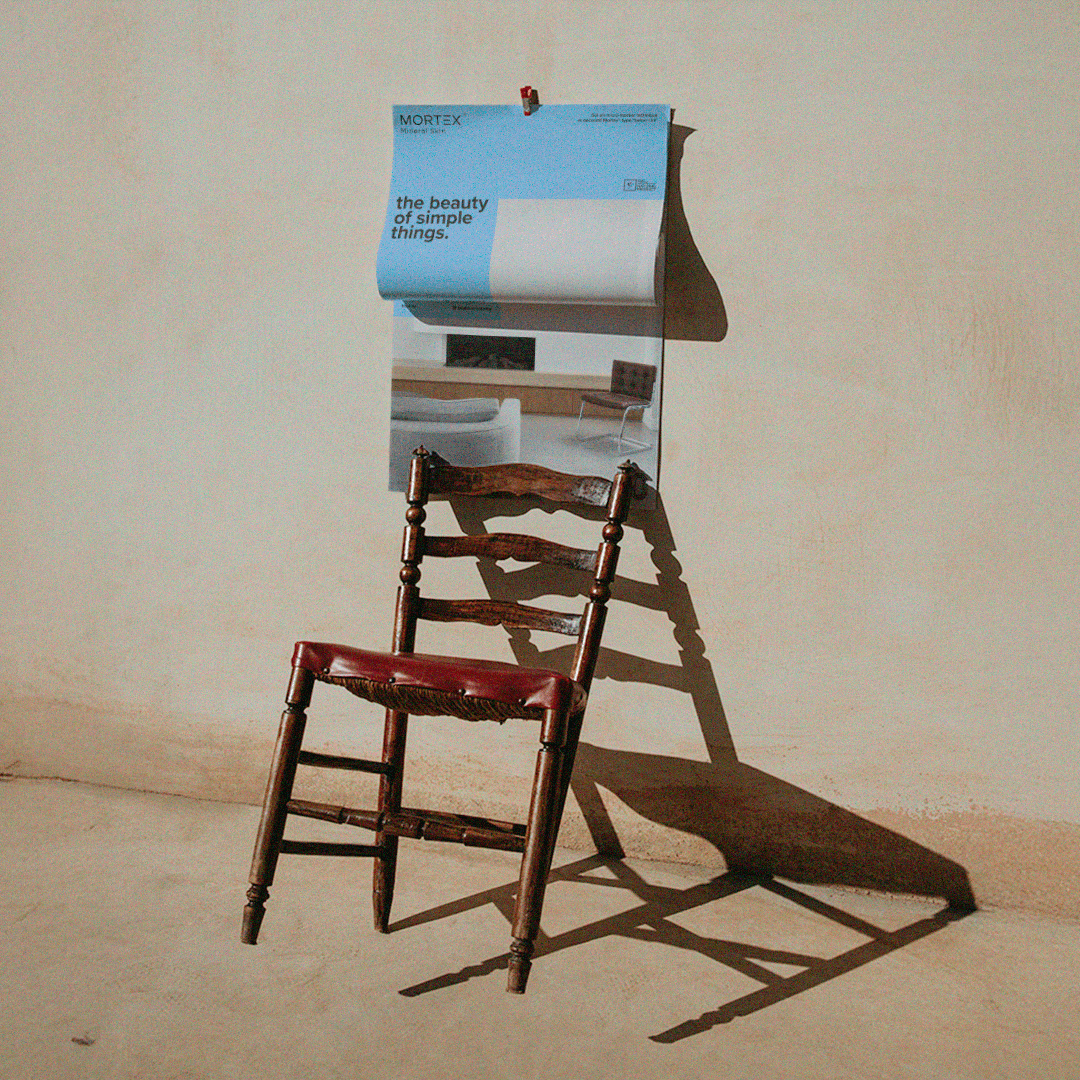 a minimal, sunlit interior scene with an old wooden chair casting a long shadow against a textured wall, above which hangs a blue-toned poster reading “the beauty of simple things.” The warm light and empty space create a quiet, contemplative, almost poetic atmosphere.
