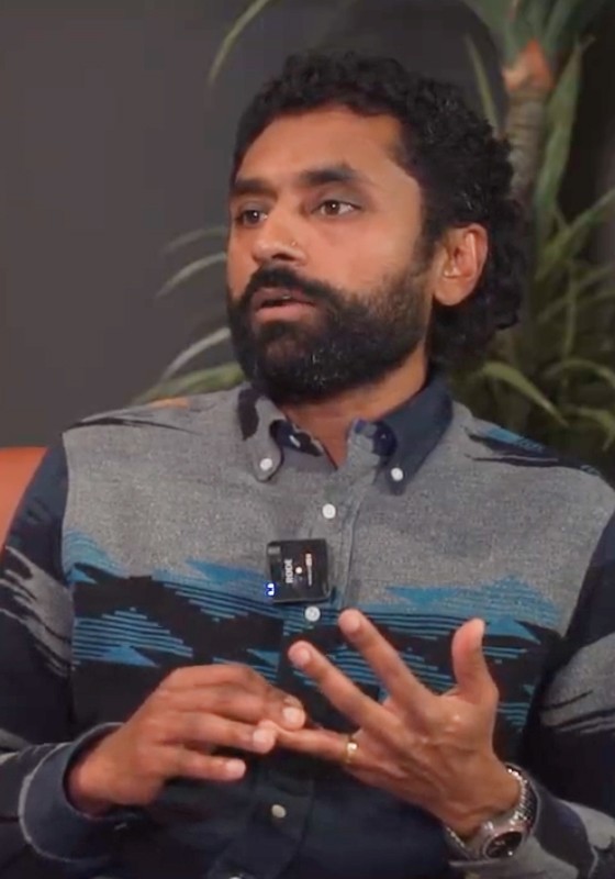 Man in a dark sweater and woman in glasses wearing a navy blouse, sitting together at a desk with a laptop and smiling.