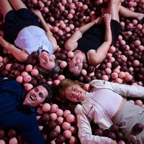Four people lying on their backs in a ball pit filled with brown and pink balls, smiling and laughing.