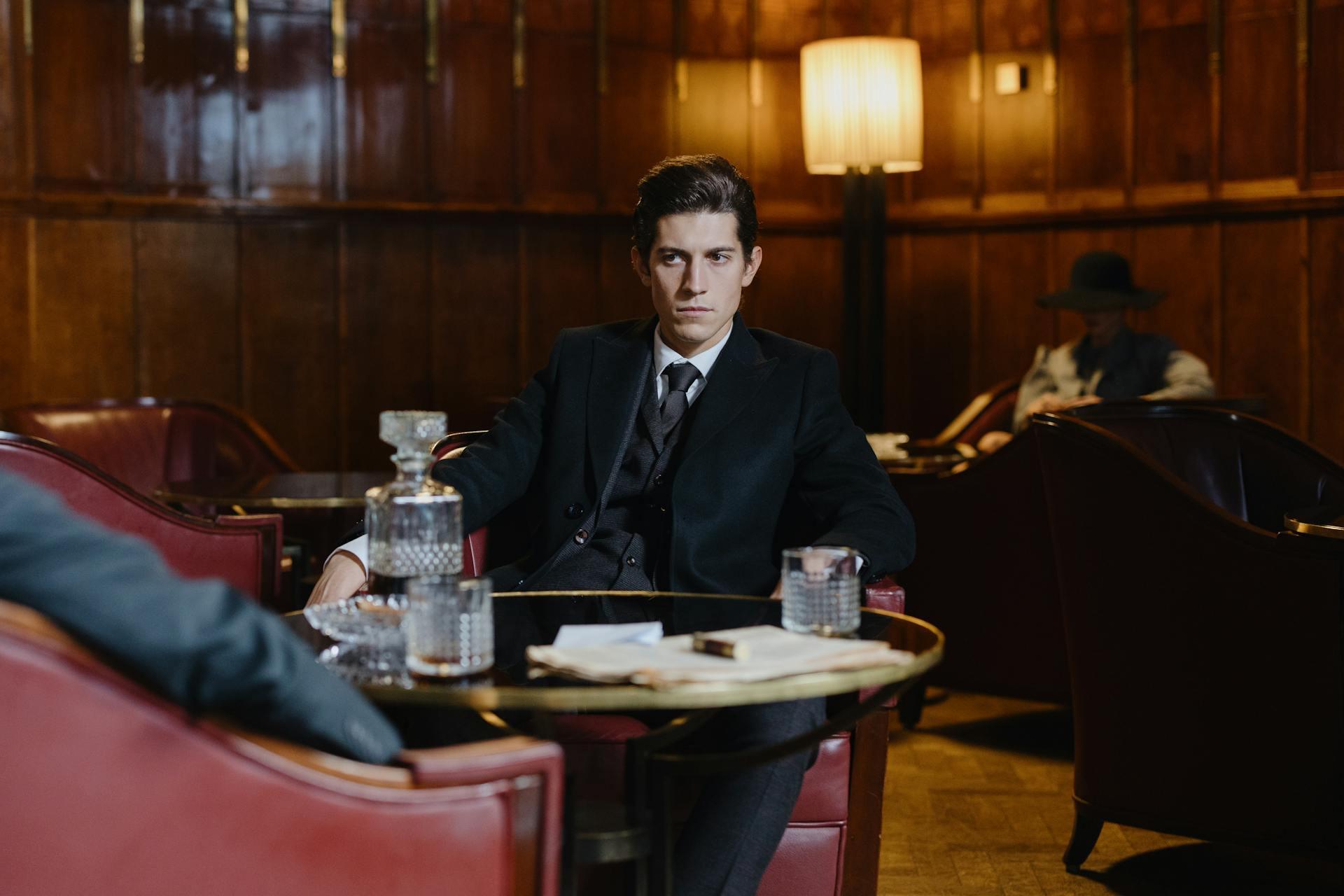 A man in a black three-piece suit sitting in a dimly lit, wood-paneled club, looking intently across a small round table set with a glass decanter and tumbler.