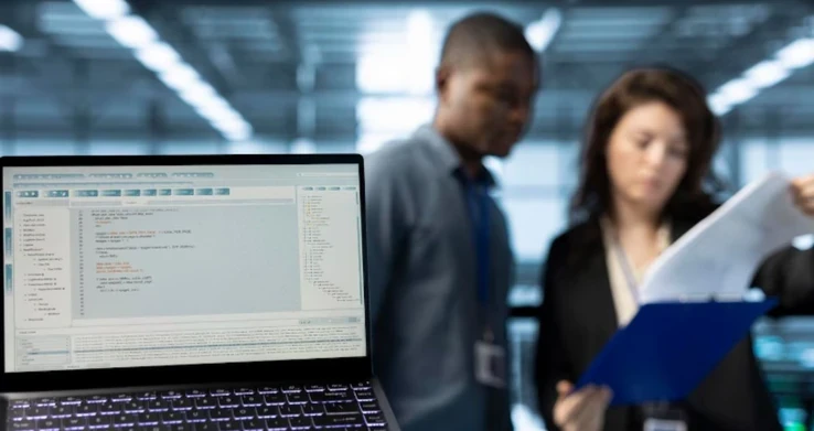 Laptop exibindo código sobre uma mesa, com colegas conversando ao fundo