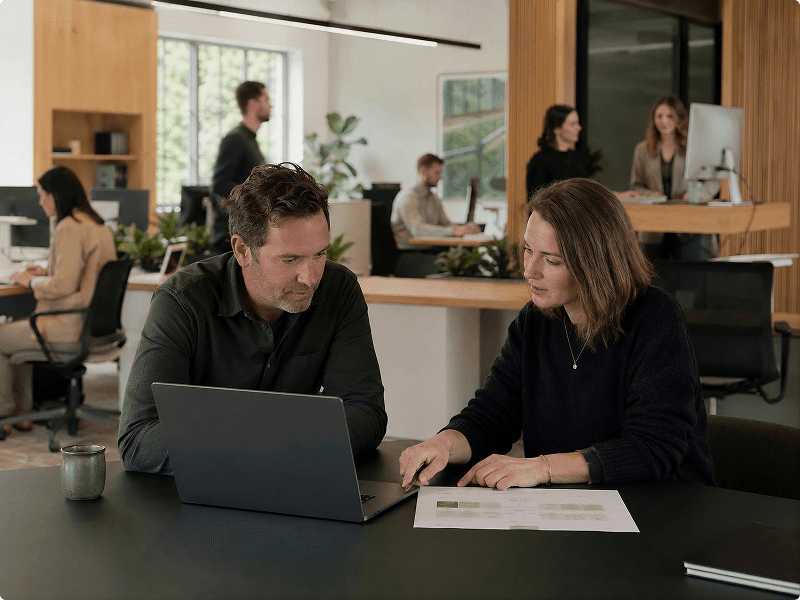 Two coworkers discuss a project at a modern office table, with other employees in the background.