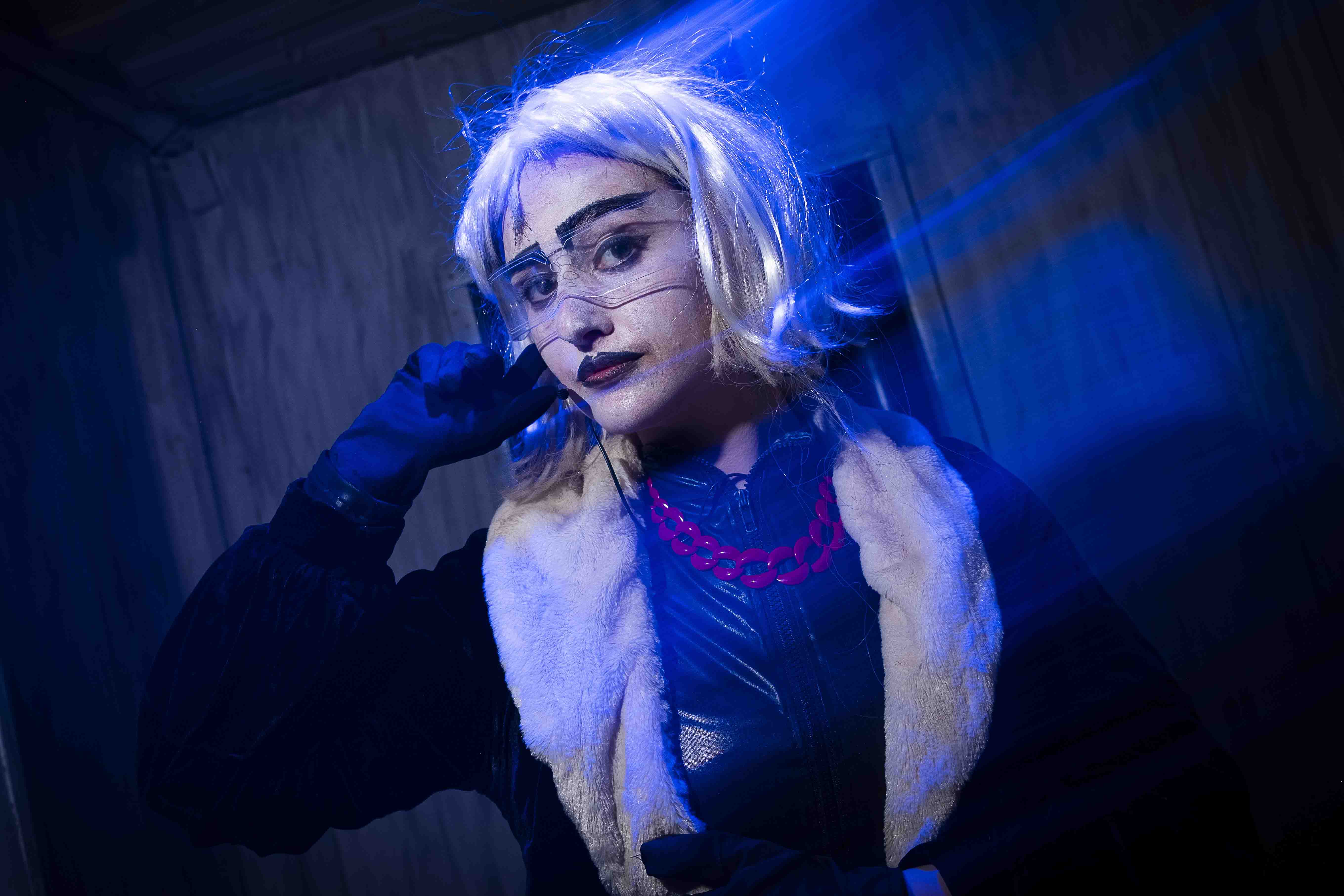 Cosplayer in a dark, dramatic pose.  They have platinum blonde hair, dark makeup, and are wearing a fur-trimmed coat and stylish accessories.