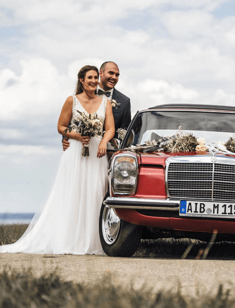 Brautpaar lehnt an rotem Oldtimer während Hochzeitsshooting im Freien