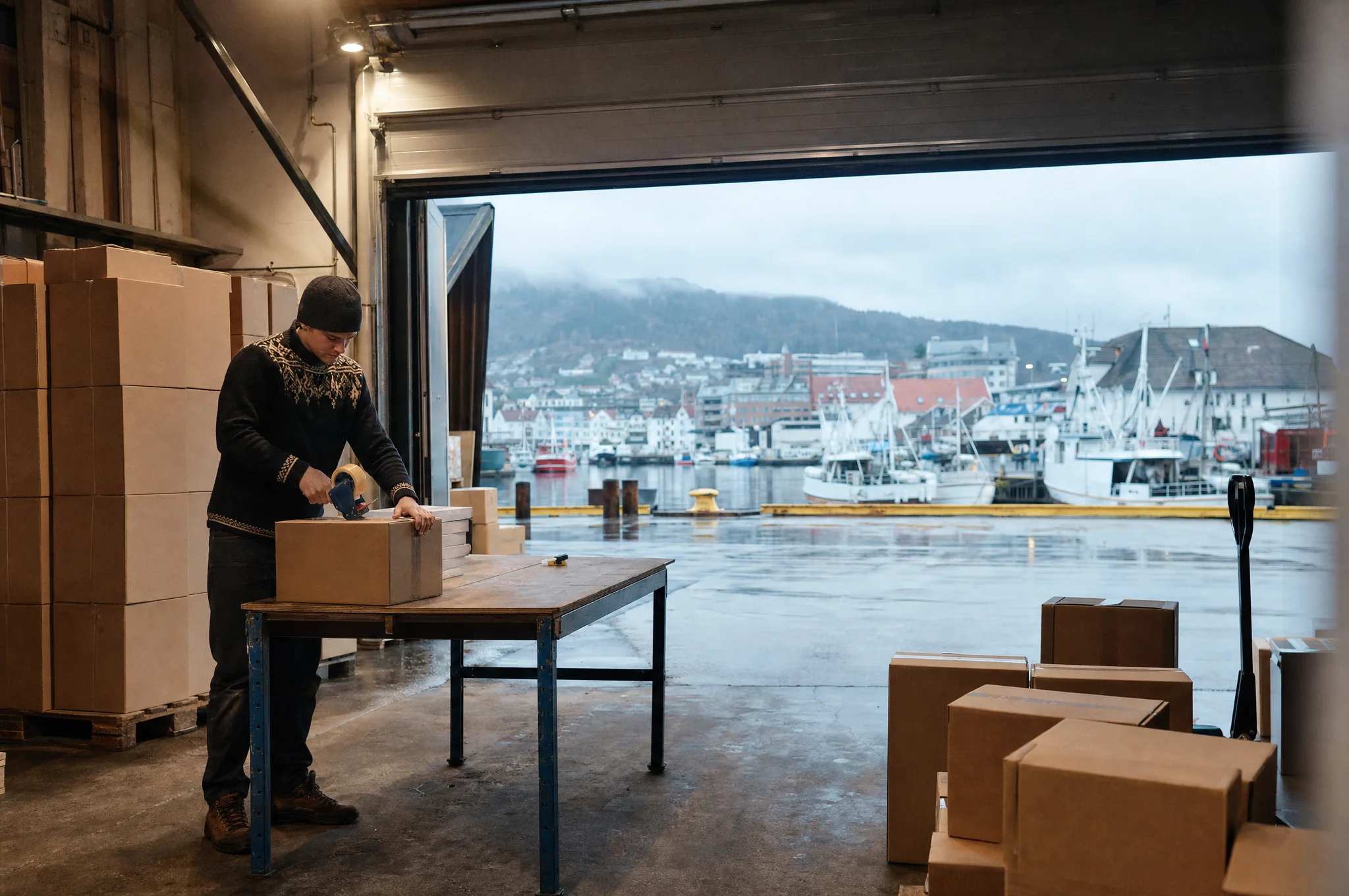 A man taping boxes in a small warehouse on a rainy Bergen quay