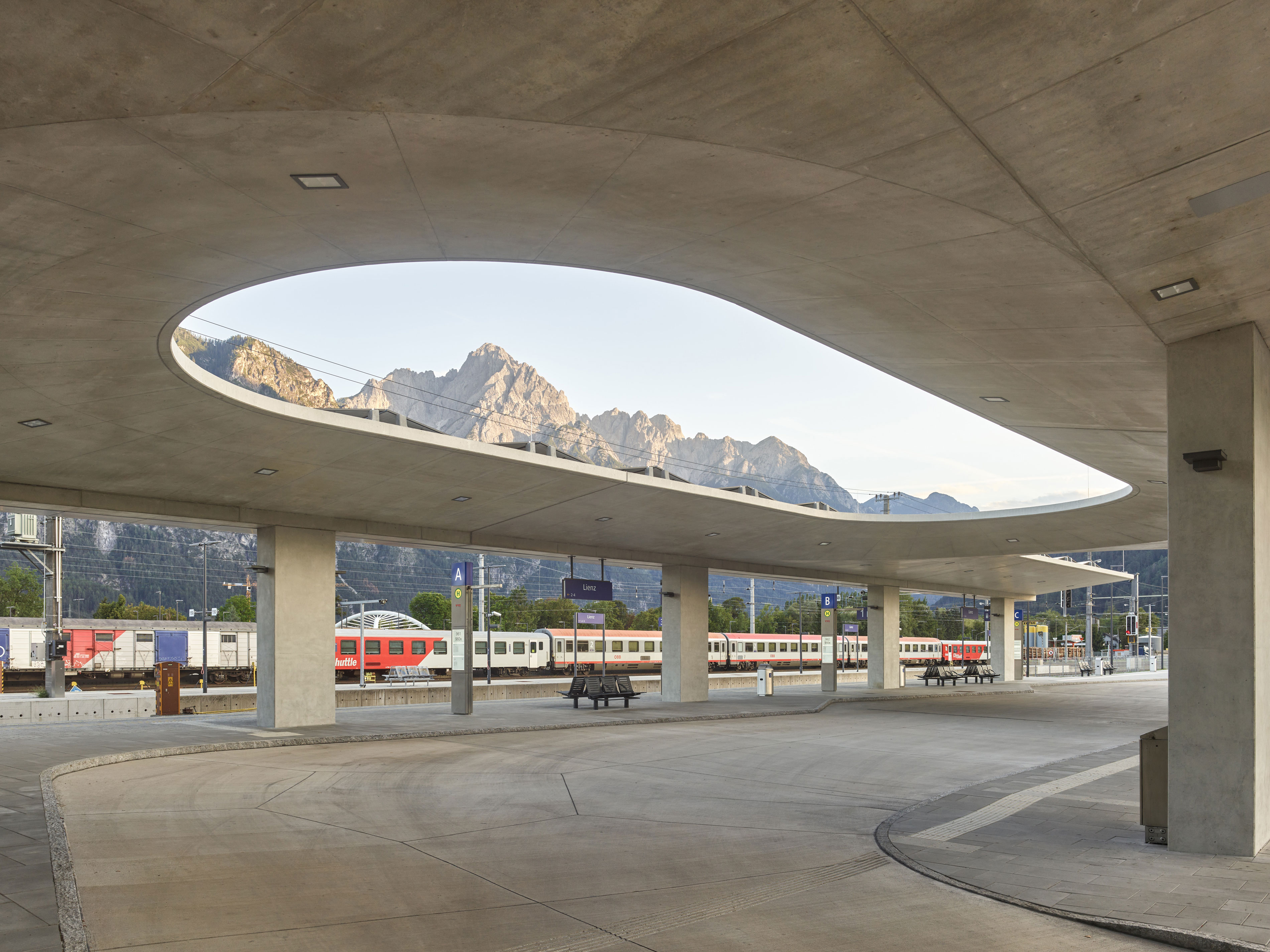 Concrete canopy with oval opening above a train platform, train waiting below, mountains in the background.