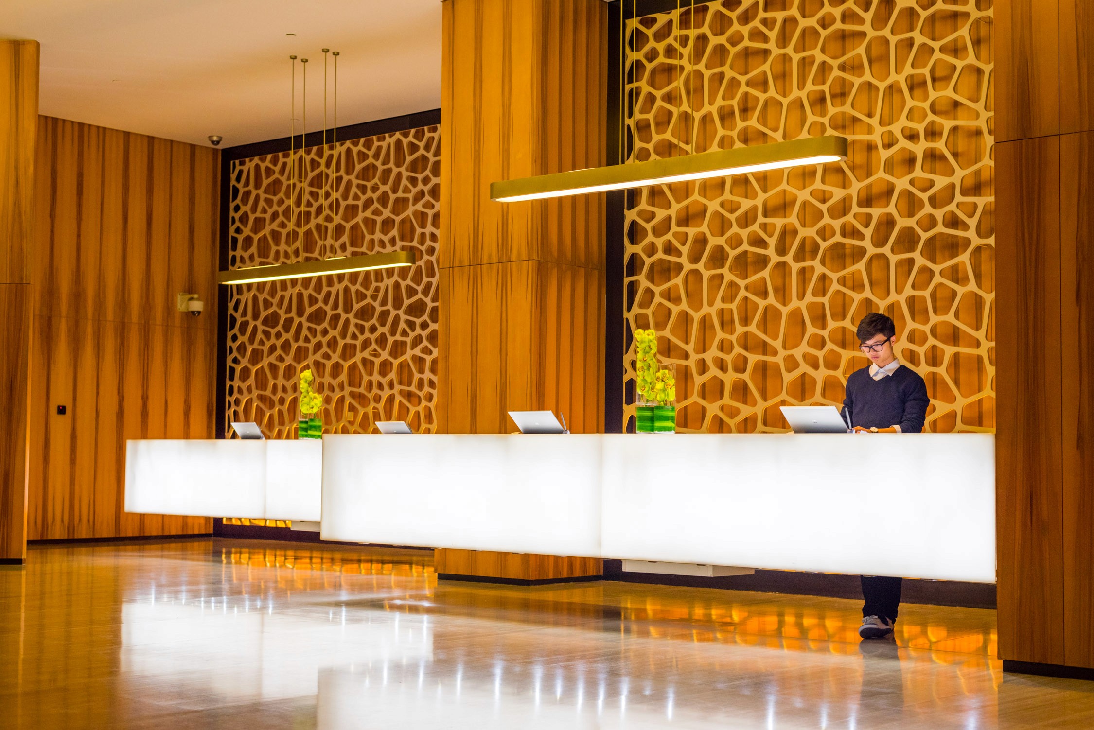 A hotel reception desk with white marble tops and gold textured walls features a staff member in a mask behind it.