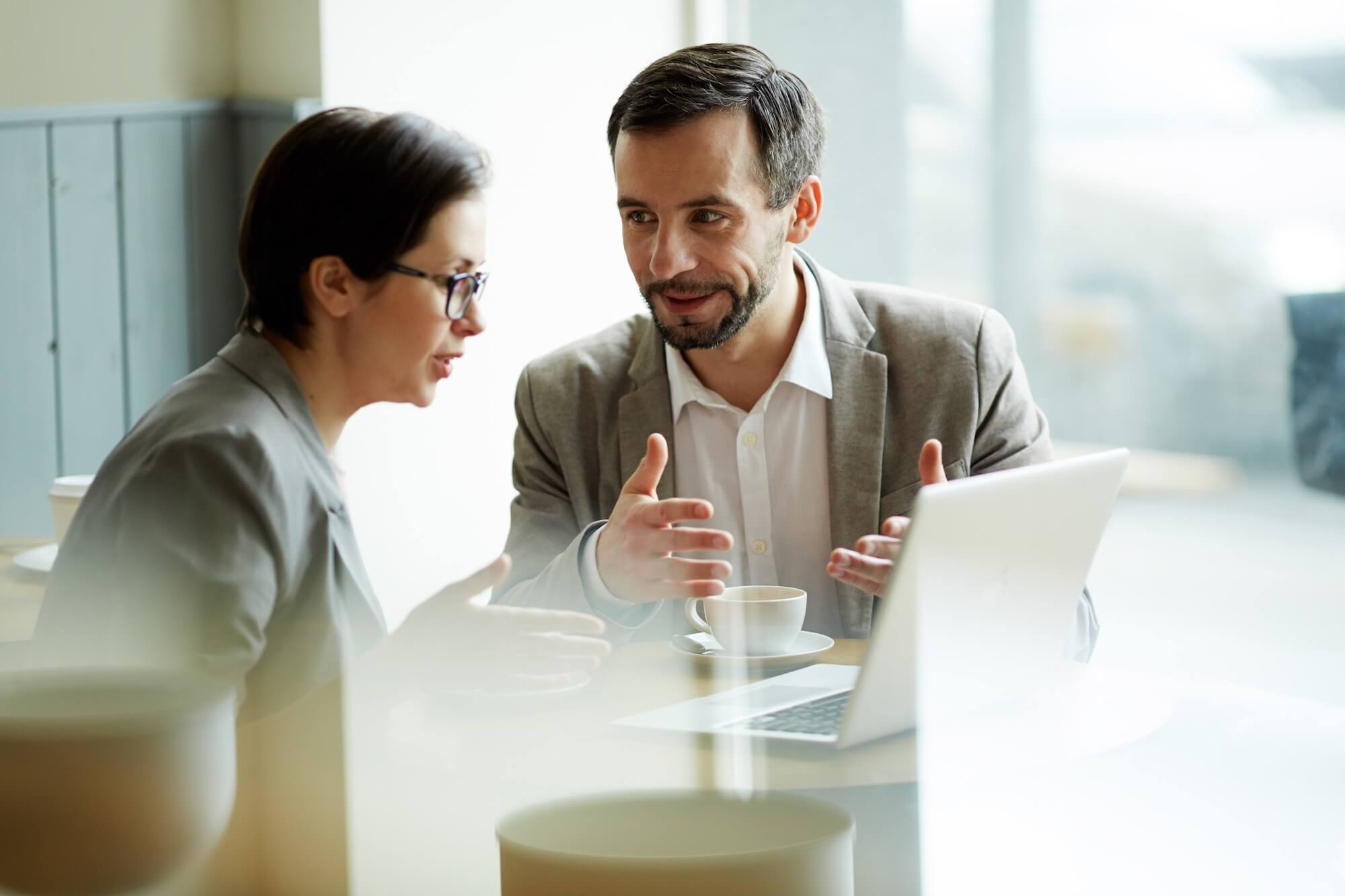 a man and a woman sitting in an office in front of a laptop, the man appears to be explaining something and has his hands in front of his body