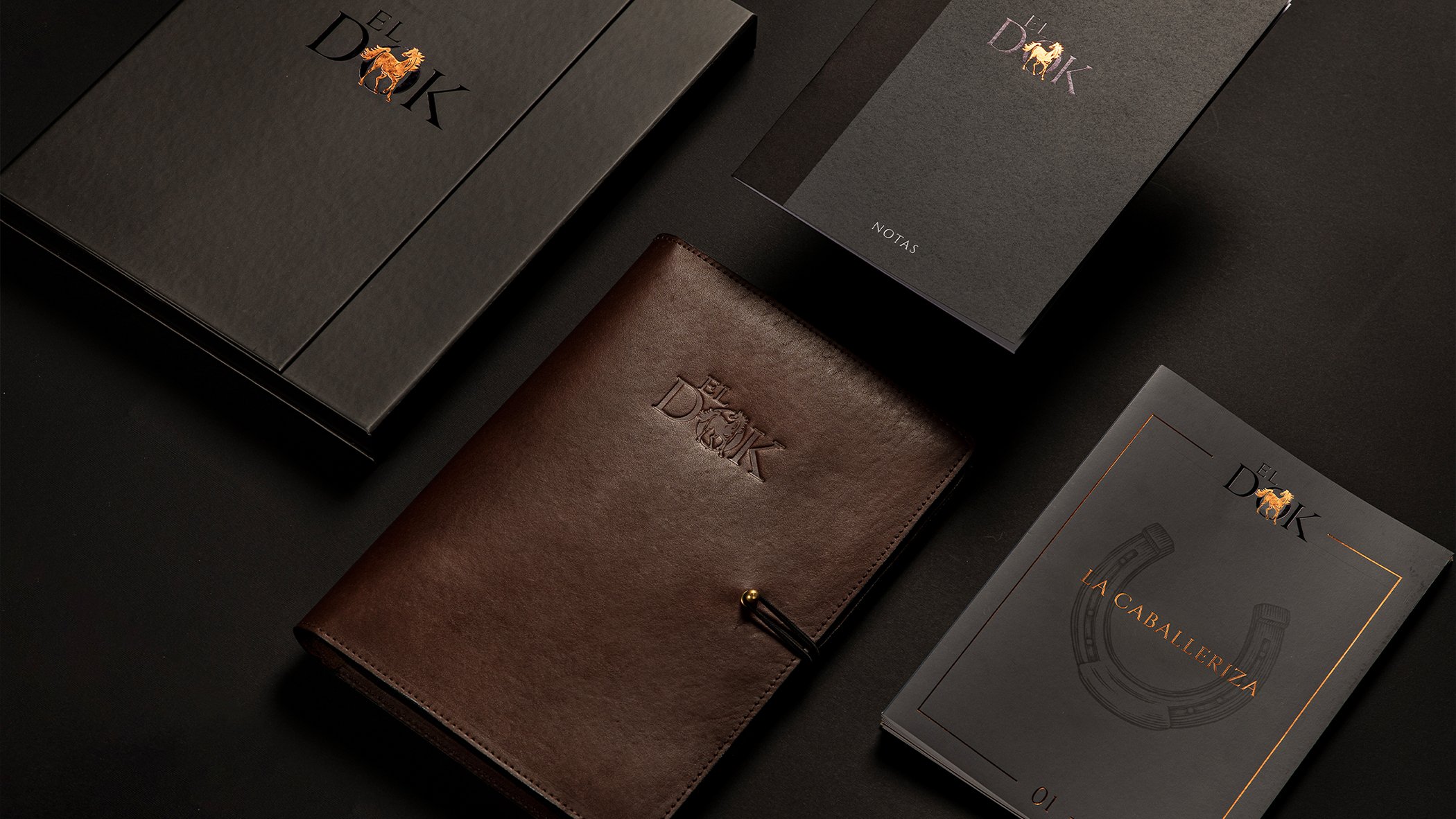 A collection of elegant, branded notebooks displayed on a dark surface. Includes a brown leather-bound book and black notebooks with gold and orange accents.