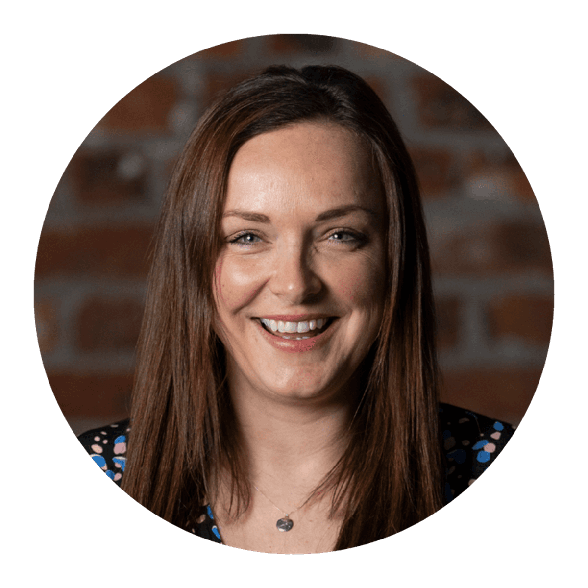 A circular portrait of a person with long brown hair, wearing a patterned top and a small necklace, posed against a brick wall background.