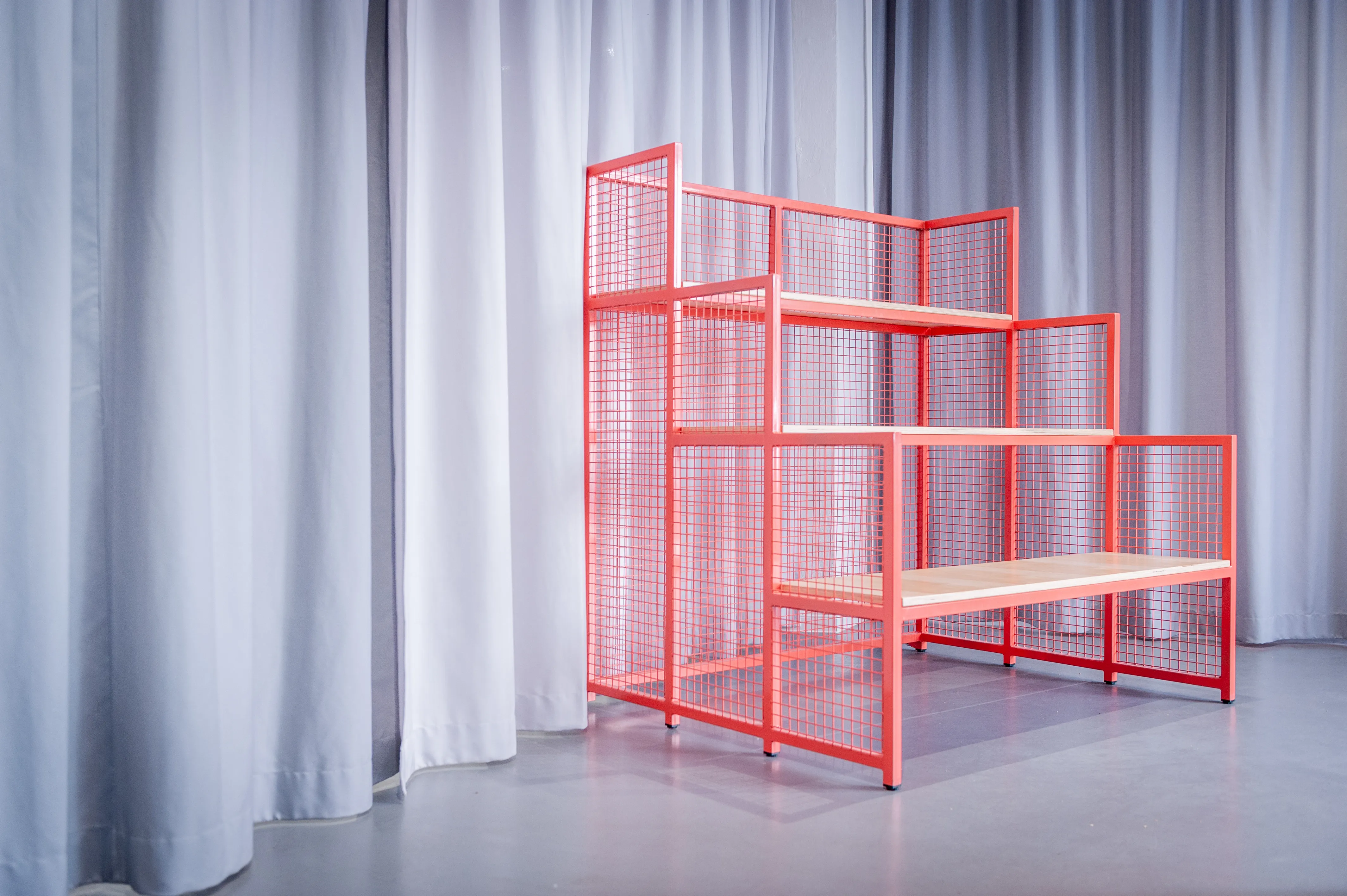 Stepped shelf module in coral-red wire mesh with wooden platforms at HEY CONTENT STUDIO, standing in front of grey curtains.