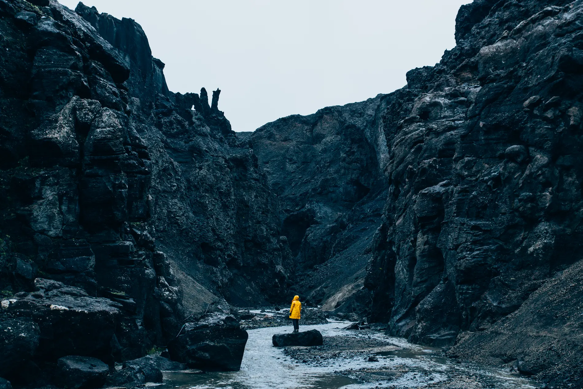 A figure in a yellow raincoat standing in a dark volcanic canyon beside a river