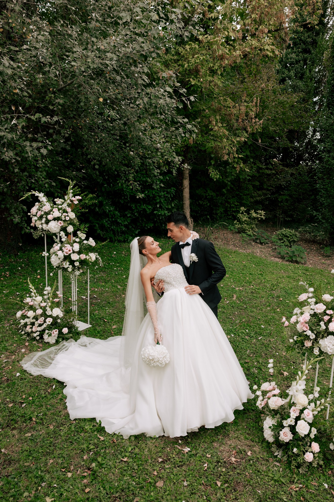 Eleonora e Fabio si abbracciano nel parco di Villa Ferrari, circondati da fiori bianchi e verde