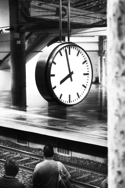 Black and white photo capturing a large clock at a railway station with people waiting on the platform.
