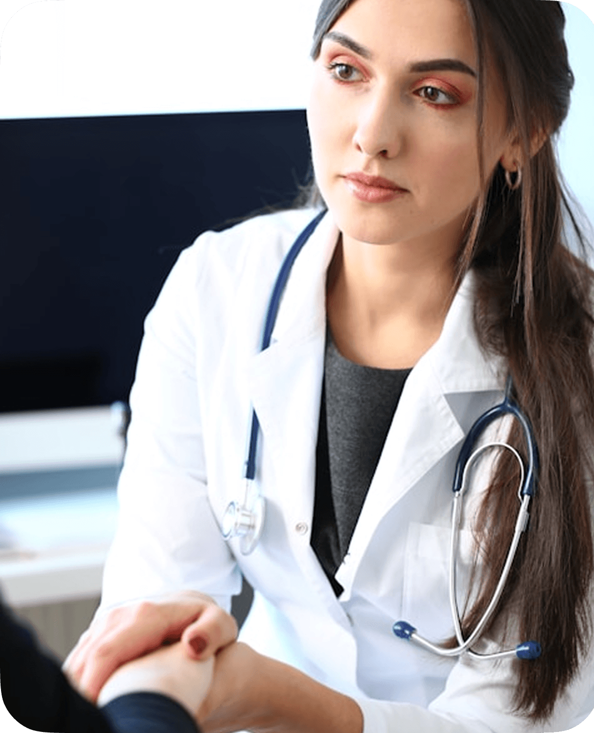 A female doctor with long brown hair, wearing a white coat and stethoscope, attentively listens while holding a patient's hand in a modern office setting.