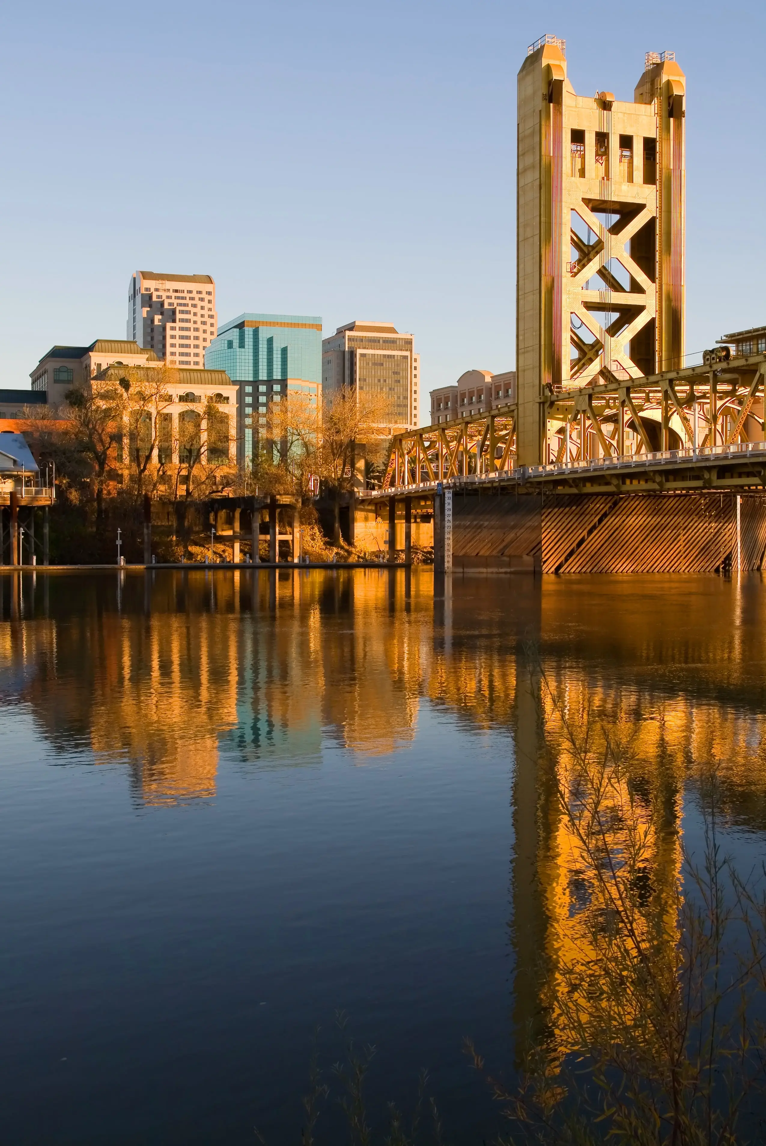 A bridge with yellow towers spans a calm river, with city buildings in the background reflecting in the water at sunset.