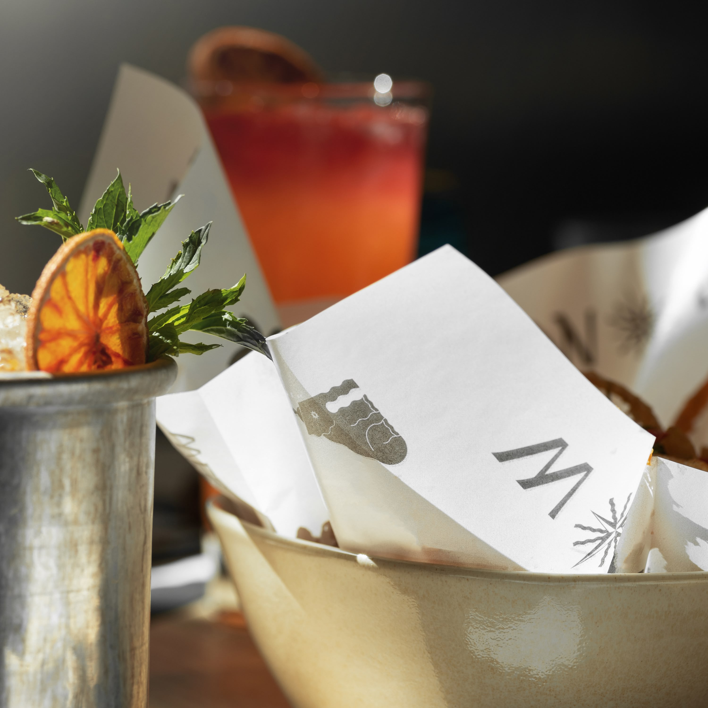 East by West restaurant bar setup with orange-garnished cocktail glass, mint sprig, silver cup, branded paper napkin basket, and ombre sunset-hued drink on wooden surface.