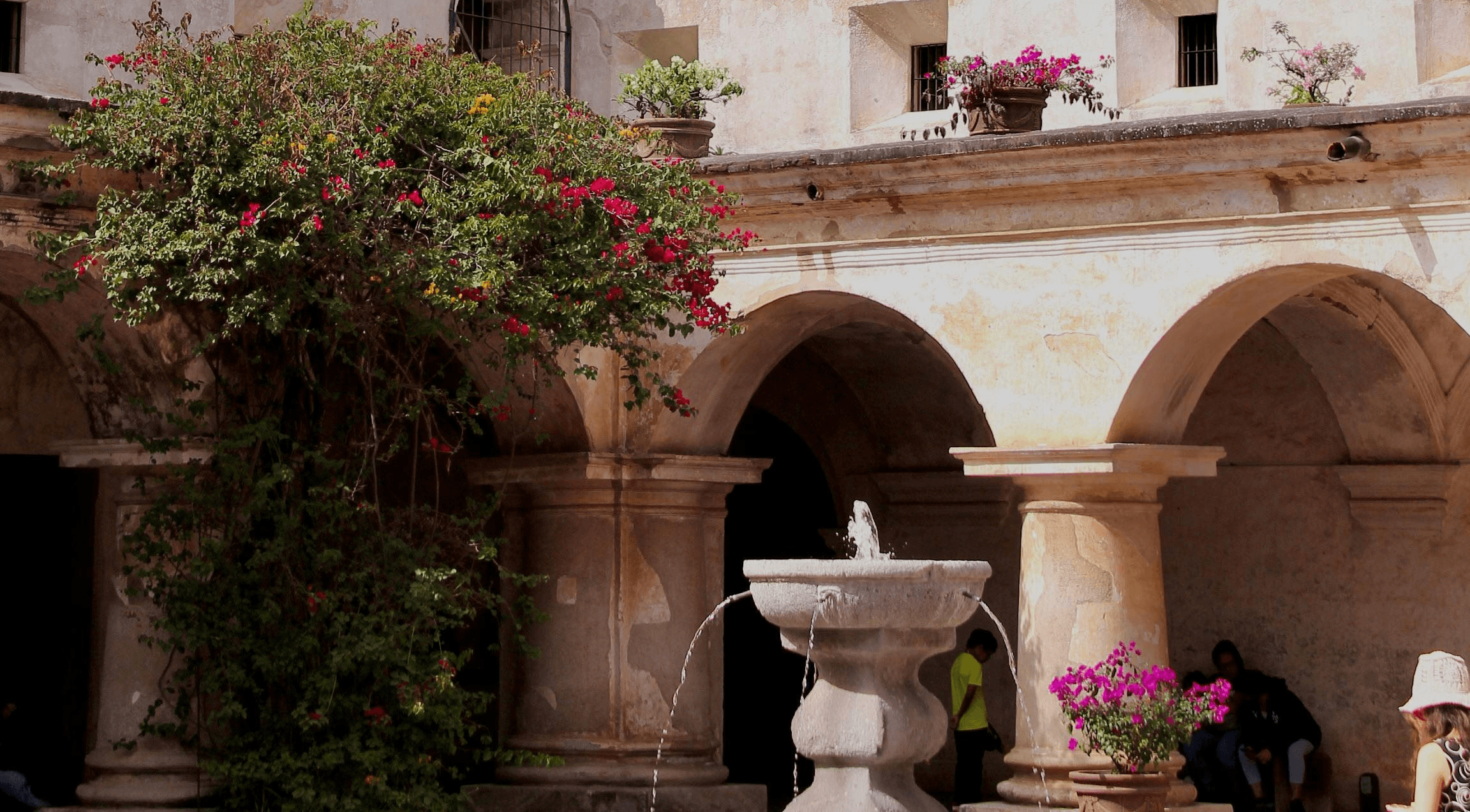 A sunlit, historic courtyard features a rustic stone fountain with water flowing gently, surrounded by arches and vibrant potted flowers, creating a tranquil, picturesque scene.
