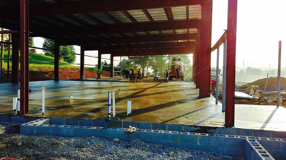Concrete foundation with reflected sunlight with red metal support structure in progress of being erected and surrounded by gravel and construction equipment.