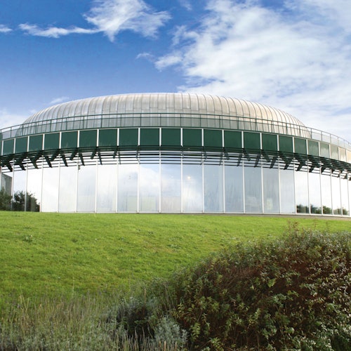 Ein kuppelförmiges, gläsernes Gebäude mit einer grünen Basis, vor einem wolkigen blauen Himmel, umgeben von grünem Gras und Sträuchern.