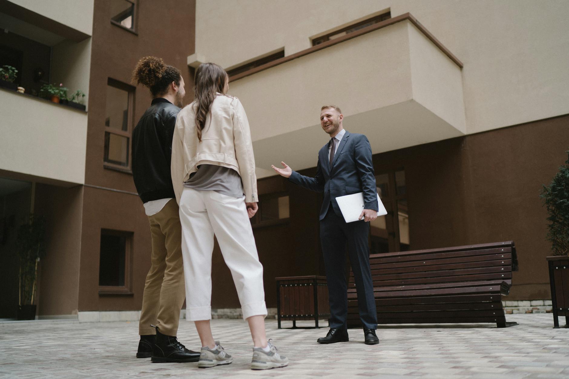 Estate agent in suit gesturing towards a brick apartment block while speaking to a couple in an outdoor courtyard.