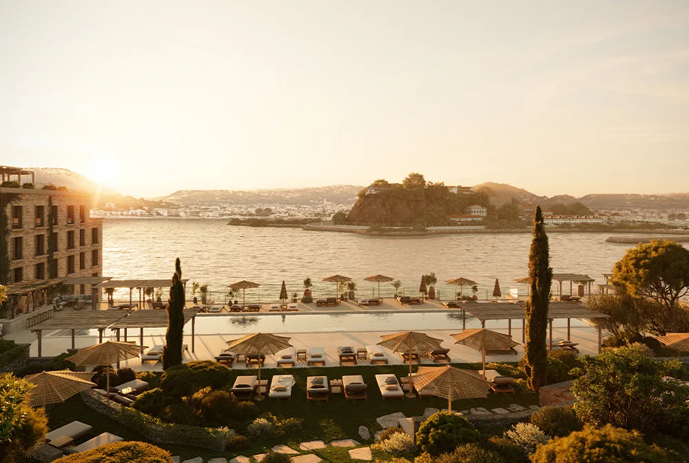 Weitläufige Hotelterrasse mit Infinity-Pool und zahlreichen Lounge-Inseln unter Bast-Sonnenschirmen. Im Hintergrund erstreckt sich eine malerische Küstenstadt am Meer während der goldenen Stunde.