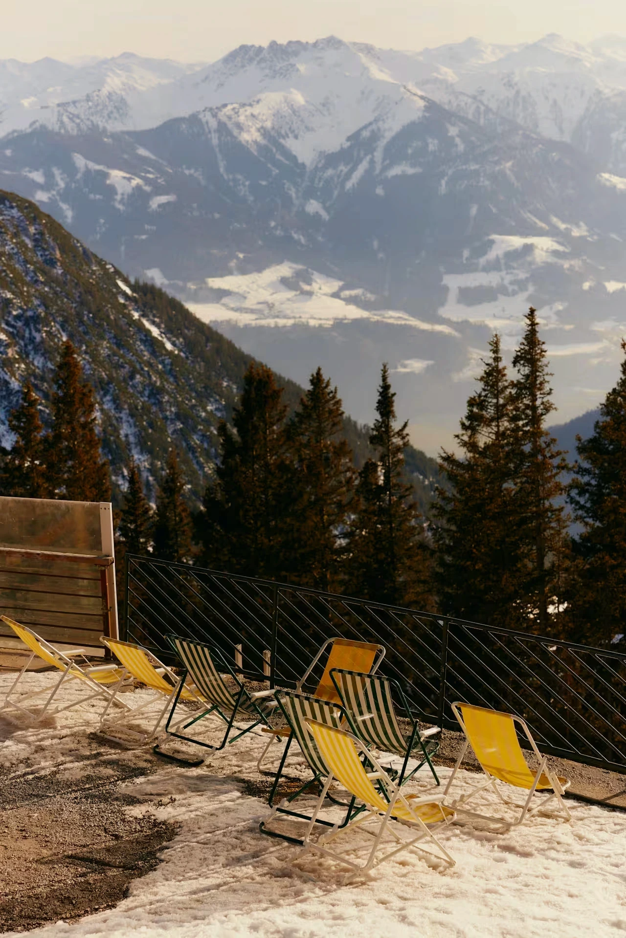 Empty deck chairs lined up on a snowy mountain terrace with pine trees and snow-capped peaks behind them.