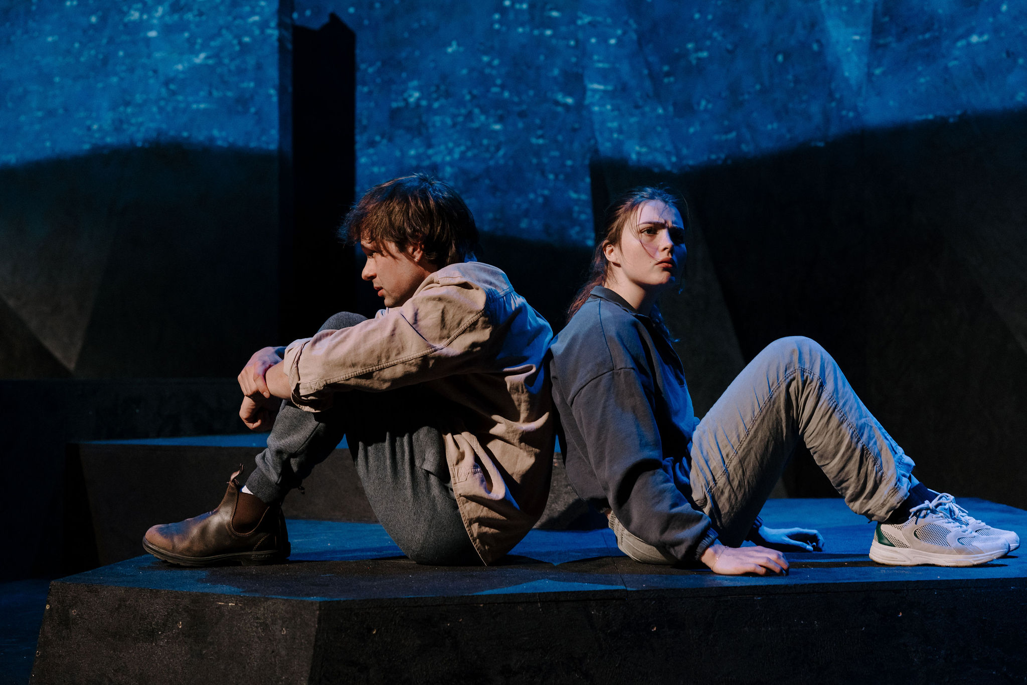 Two performers sit on a raised platform under deep blue lighting; one rests against the other while the second gazes upward.