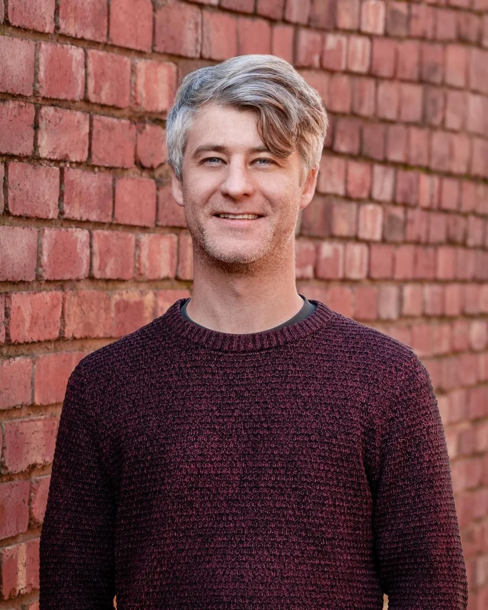 Man with gray hair wearing a burgundy sweater standing against a brick wall.