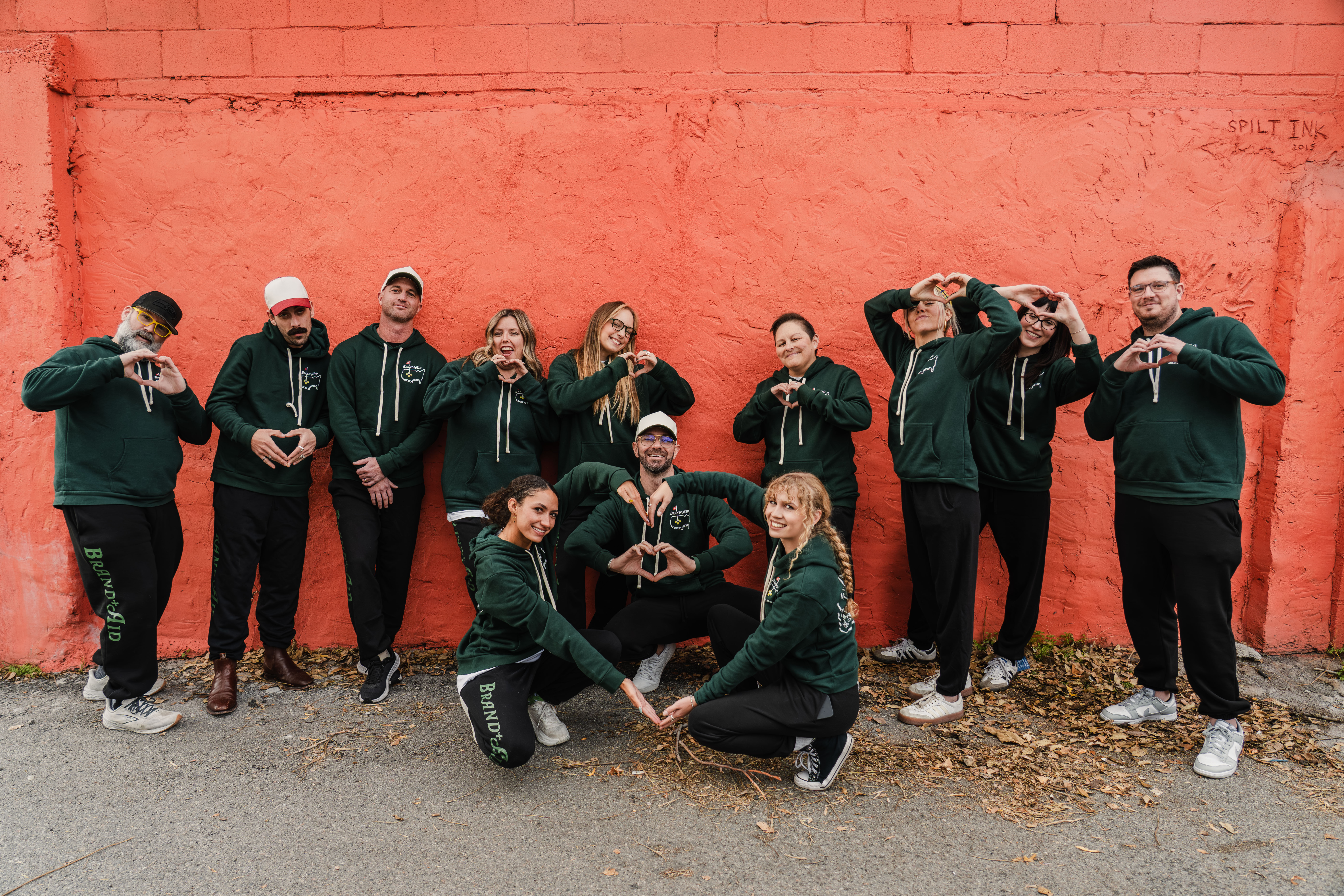 The brand aid team in matching track suits all standing in front of an orange wall. Three people are in the front forming a heart with their arms and hands.