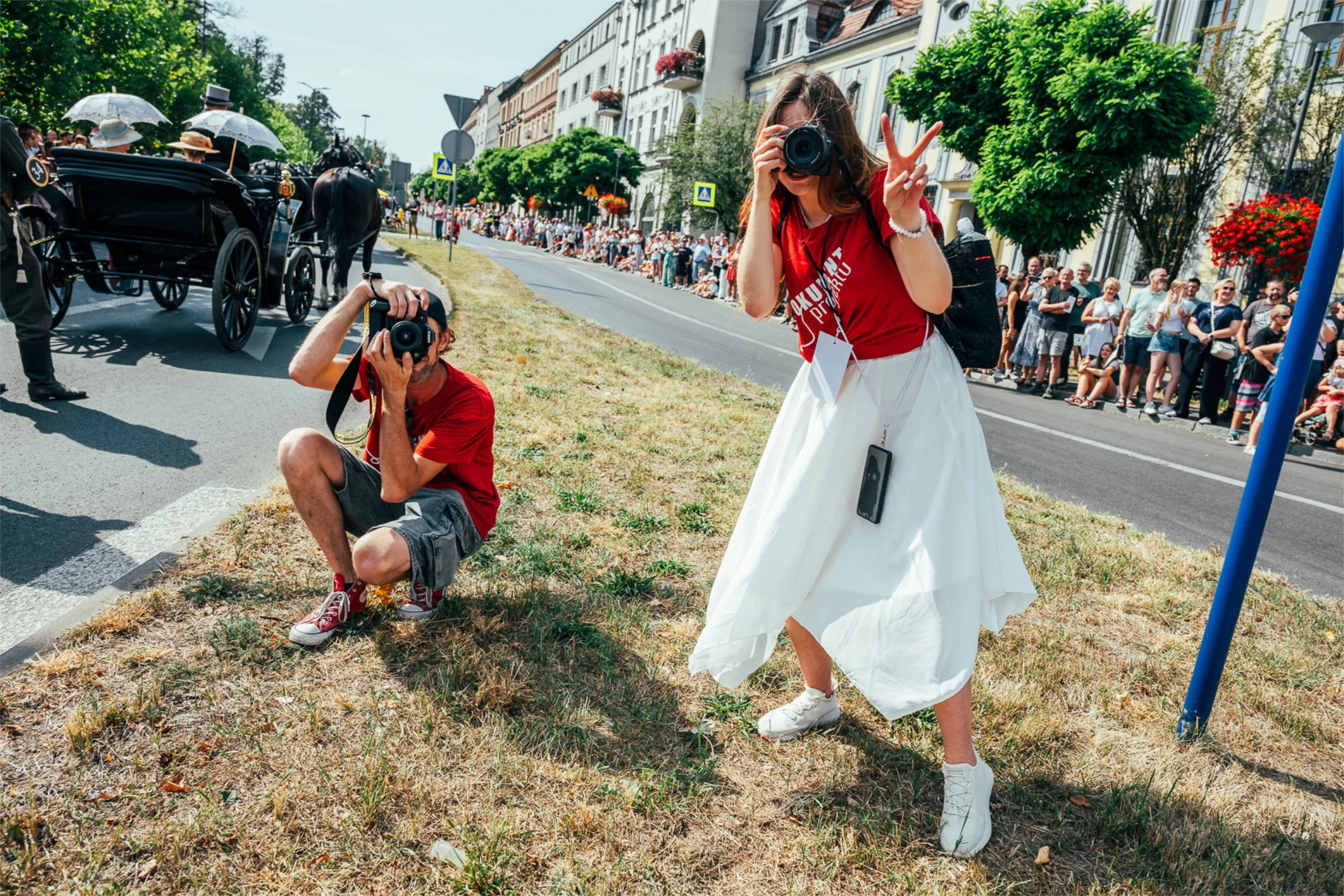 Fotografowie robią zdjęcia podczas wydarzenia. Kobieta z aparatem robi znak pokoju, obok mężczyzna w czerwonej koszulce.