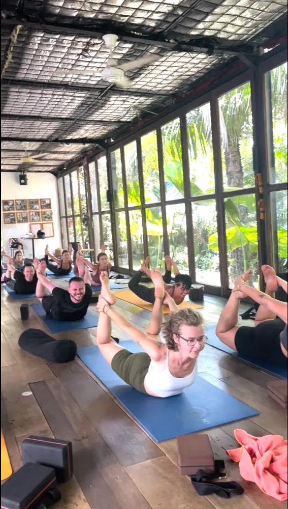 Yoga teacher training students practicing Bow Pose (Dhanurasana) together in a glass-walled studio.