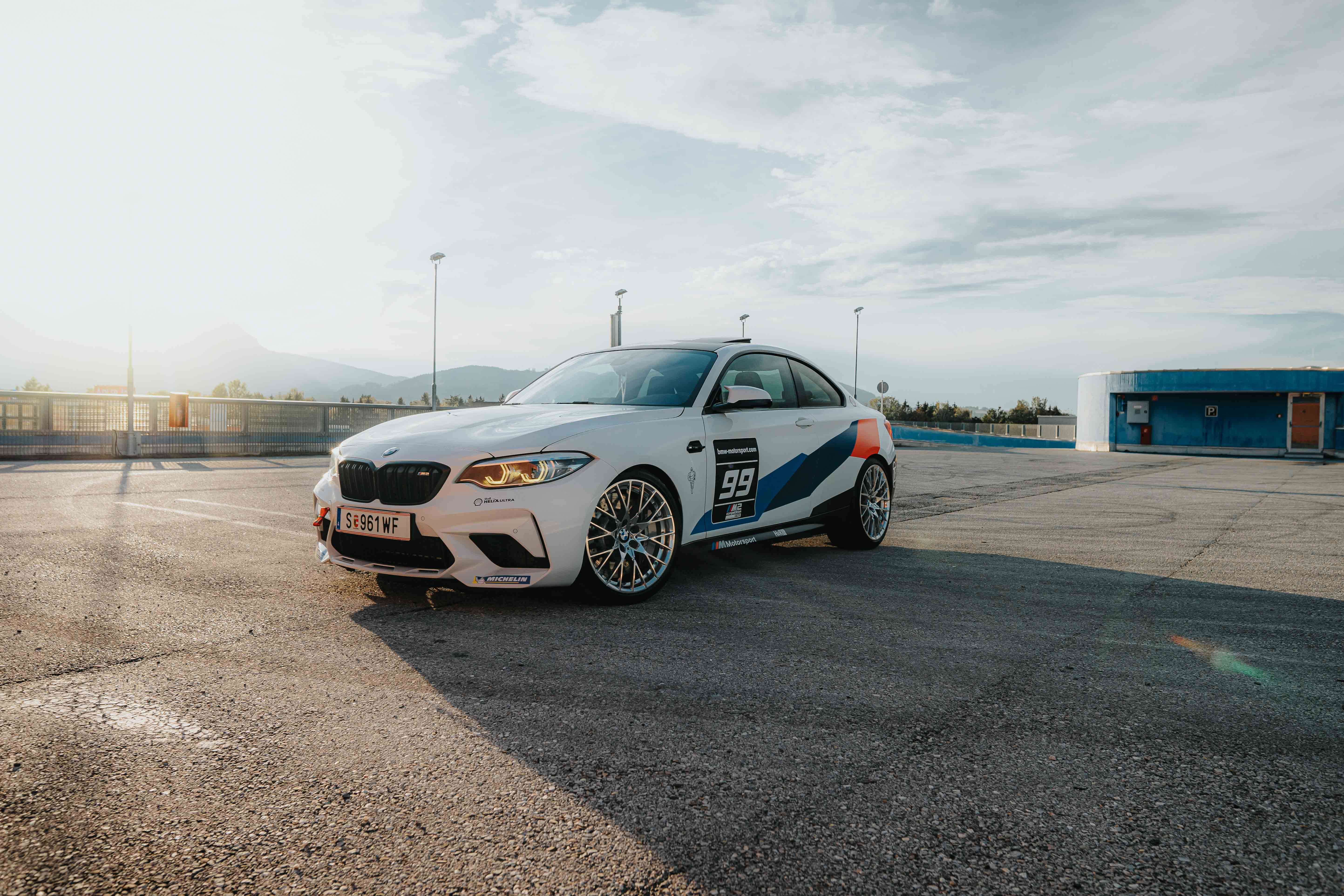 Weißer BMW M2 auf einem Parkplatz im Sonnenlicht. Der Sportwagen hat blaue und weiße Rennstreifen.