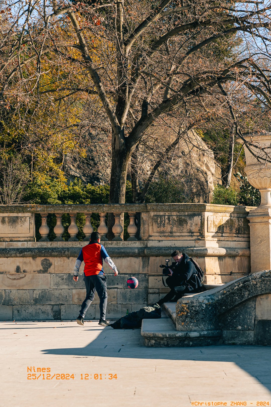 Un enfant jonglant avec un ballon sous l’objectif d’un vidéaste, dans les jardins de la Fontaine.
