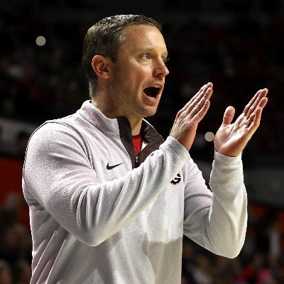 Coach Mike White wearing a light gray pullover claps his hands and shouts encouragement during a game, with a blurred crowd in the background.