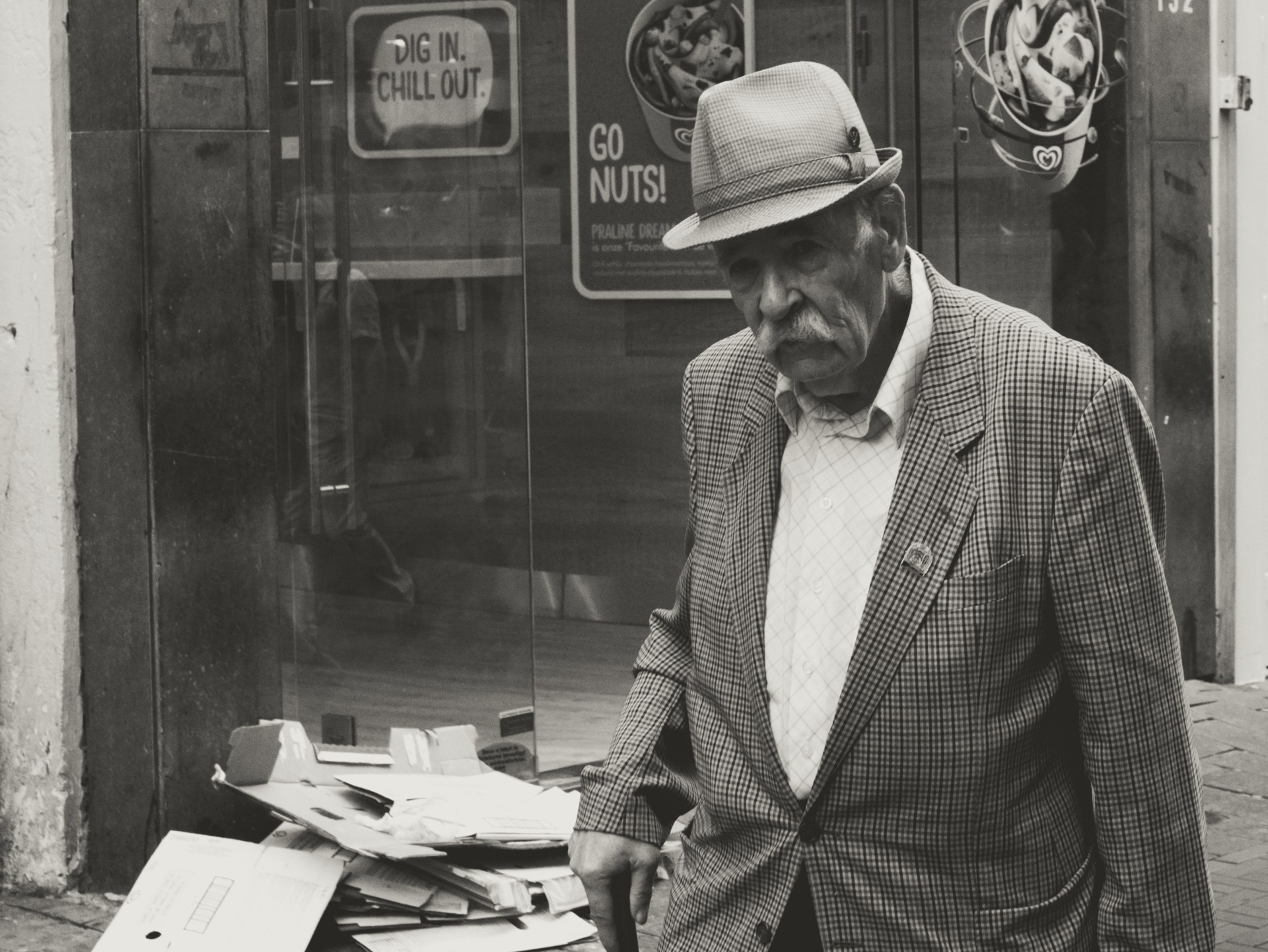 Black-and-white urban street photography of an older pedestrian by an ice cream shop window with stacked cardboard boxes, reflective storefront glass, and bold signage — ideal for content about street photography, urban life, and candid documentary images.