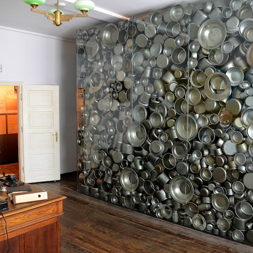 Room with a large wall display of stacked metal pots and pans, wooden desk, and open door leading to another room.