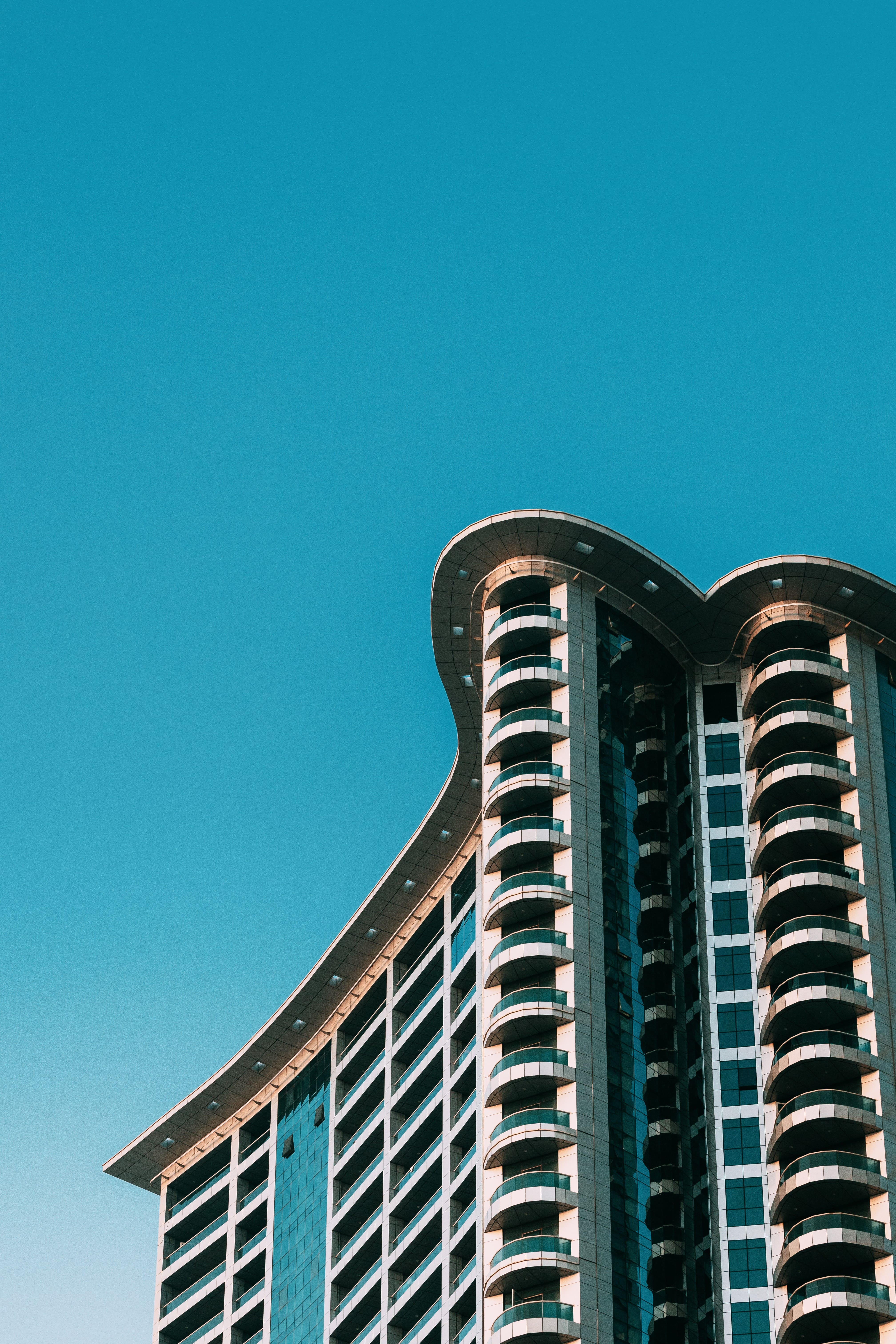 A low-angle shot looking up at a modern skyscraper with curved balconies on its upper floors, set against a vibrant, clear blue sky.