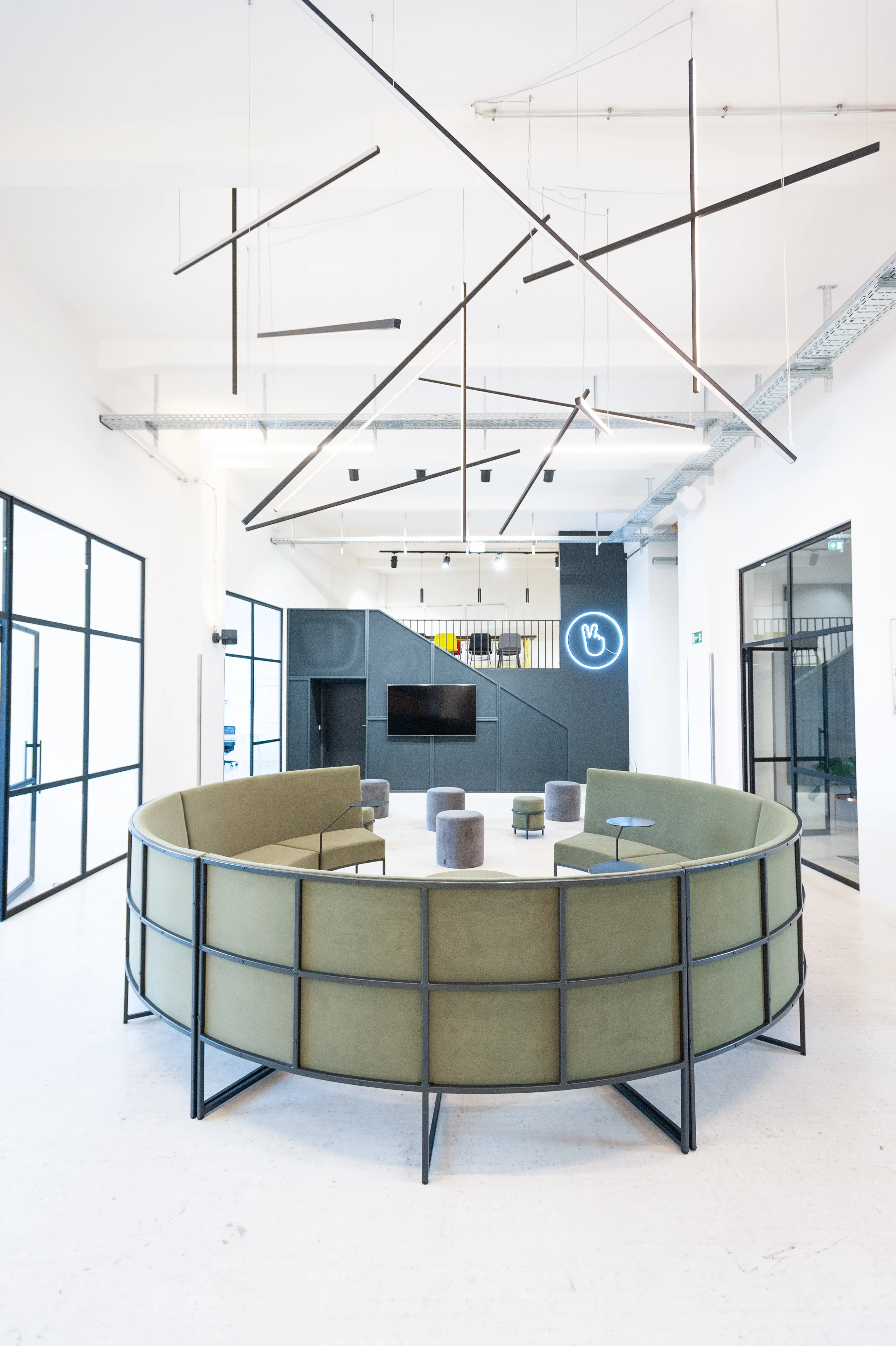 Circular sage-green lounge sofa in a black steel frame at LA RED beneath a sculptural arrangement of black linear pendant lights, with a dark kitchen block and neon hand sign in the background.