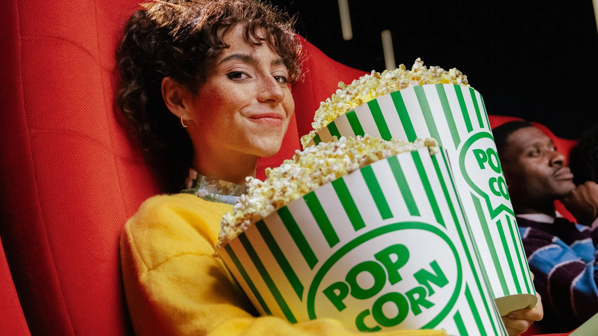 Girl with two huge popcorn boxes sitting in cinema and smiling