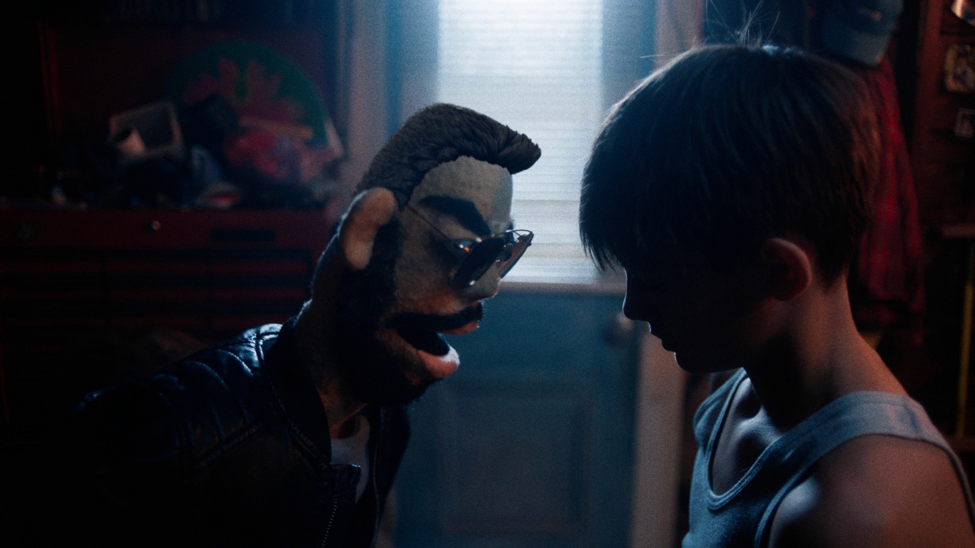 A boy in a dimly lit room faces a puppet wearing sunglasses and a leather jacket. The atmosphere is tense, with red light streaming from a window behind them.