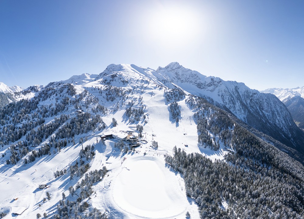 Eine atemberaubende Luftaufnahme von schneebedeckten Bergkette mit Skipisten in Hoch Oetz, umgeben von dichten Wäldern unter einem klaren blauen Himmel und hell scheinender Sonne.