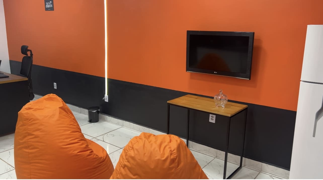 Espaço de convivência corporativo com puffs laranja, TV na parede e design moderno, promovendo conforto, criatividade e bem-estar no ambiente de trabalho.