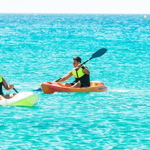 Two people kayaking on bright turquoise water, wearing yellow life vests. One is in an orange kayak, the other partly visible in a green kayak.