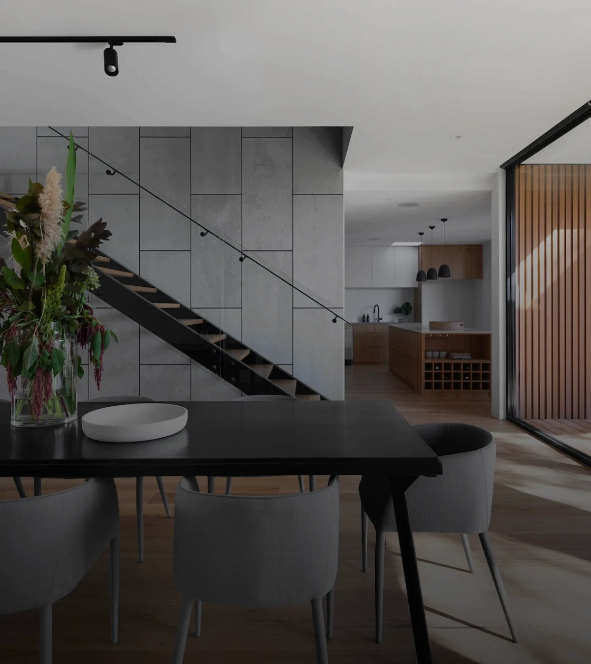 imagem de espaço residencial projetado de forma sofisticada, com tons cinza e marrons. Espaço tem uma mesa de centro com cadeiras, uma escada de fundo e outro ambiente mais atrás.