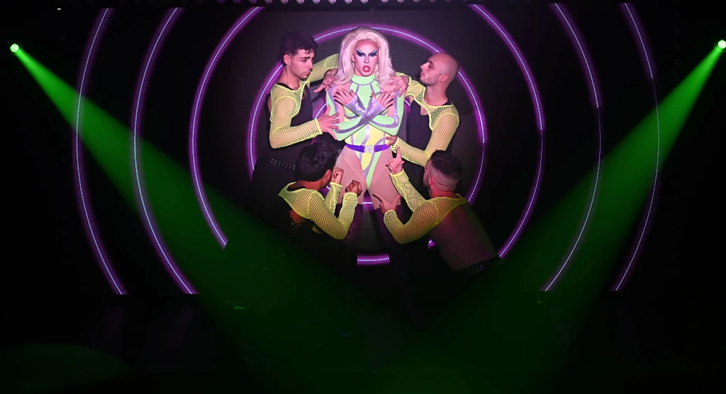 A dynamic stage shot of a central drag queen in yellow performing with backup dancers holding glowing neon hoops and vibrant background visuals.