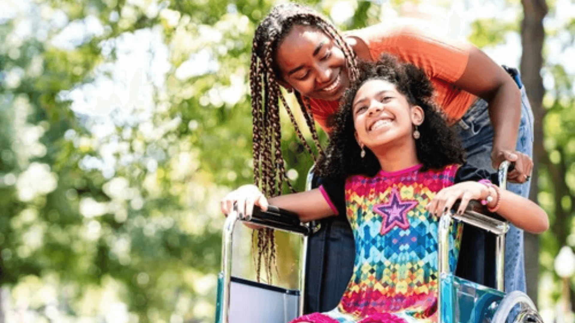 Caregiver smiles while pushing a joyful young girl in a wheelchair along a sunlit path outdoors