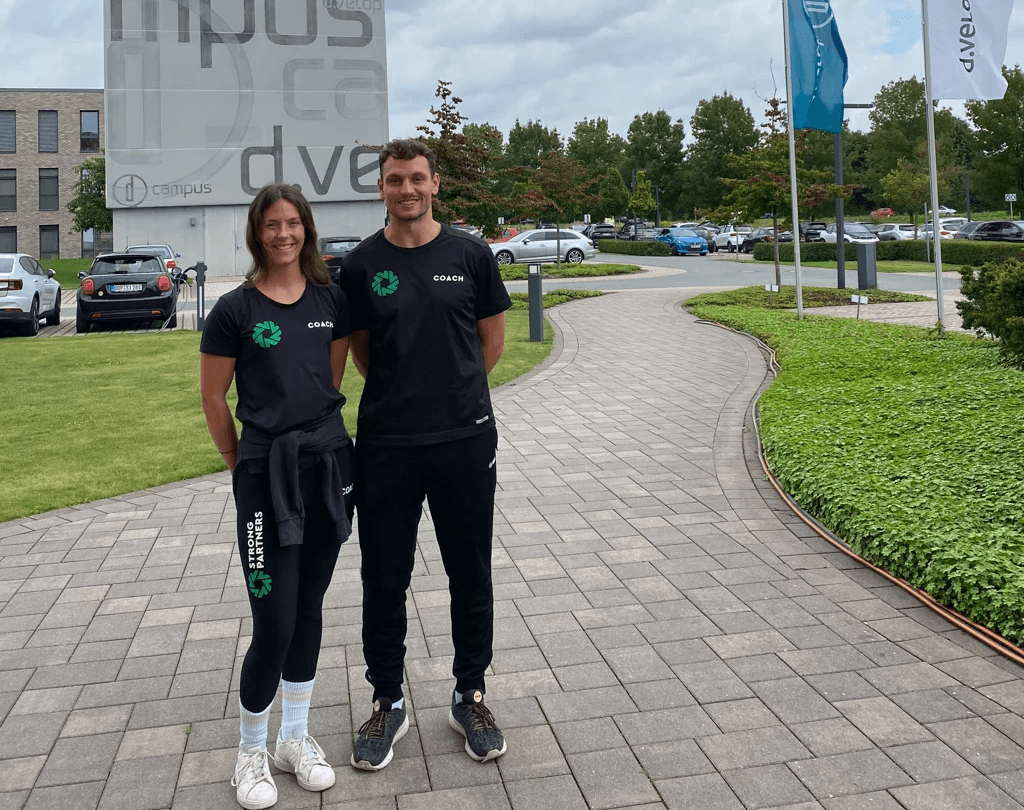 Eine junde Frau steht neben einem jungen Mann vor einem Unternehmenscampus. Beide tragen Sportkleidung von Strong Partners und lächeln in die Kamera.
